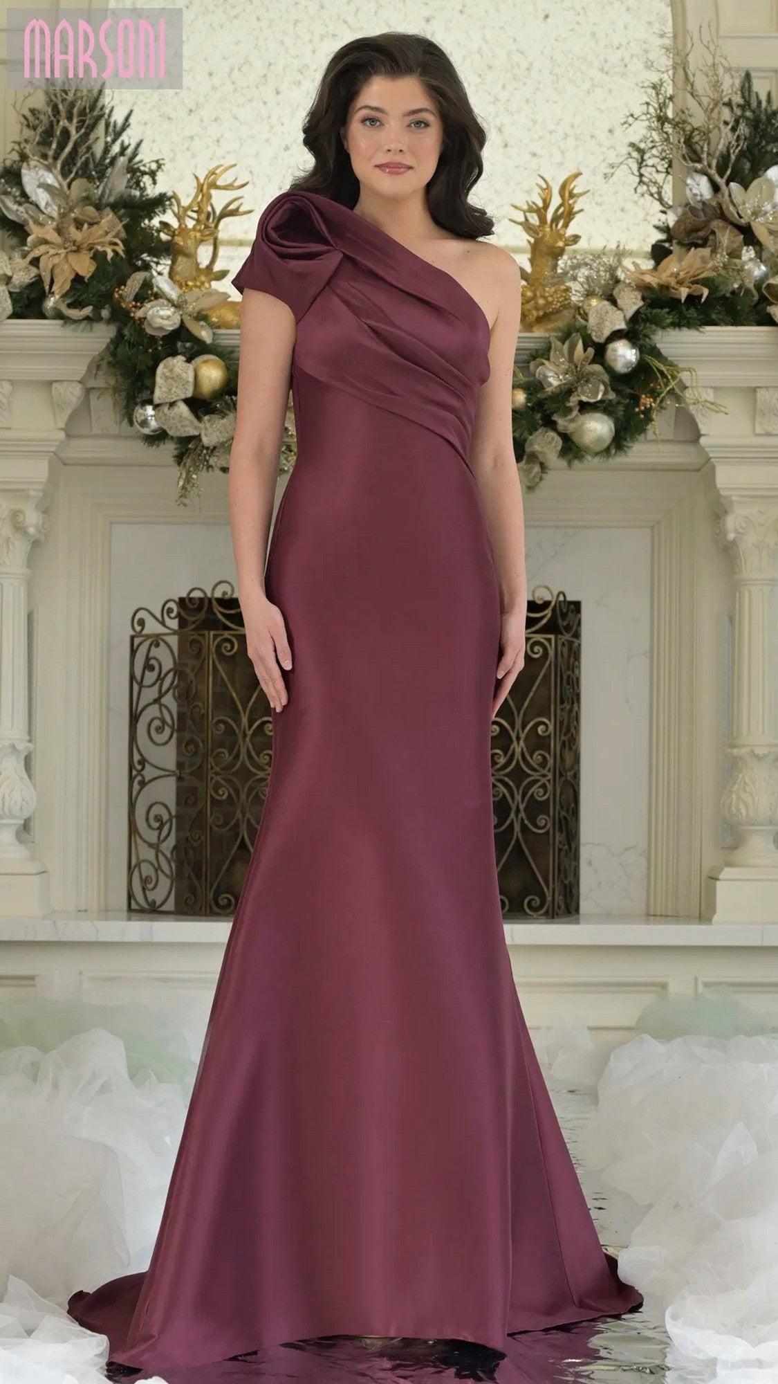 A woman in the Marsoni MV1358 long formal dress, a burgundy one-shoulder gown, stands before a mantel decorated with gold, white, and green holiday ornaments.